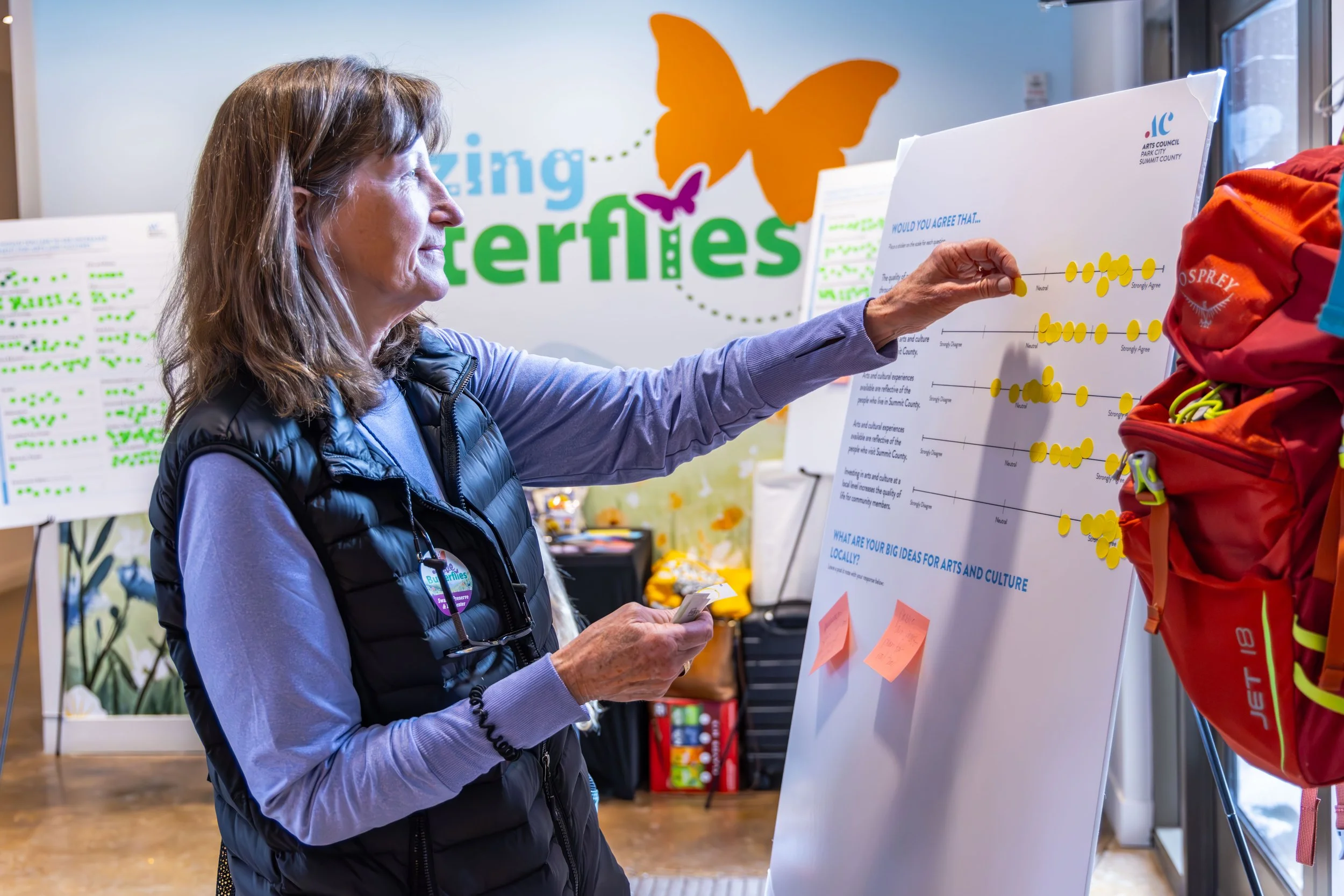 Woman placing yellow stickers on a large white board at an art and culture event, with butterfly-themed signage in the background.