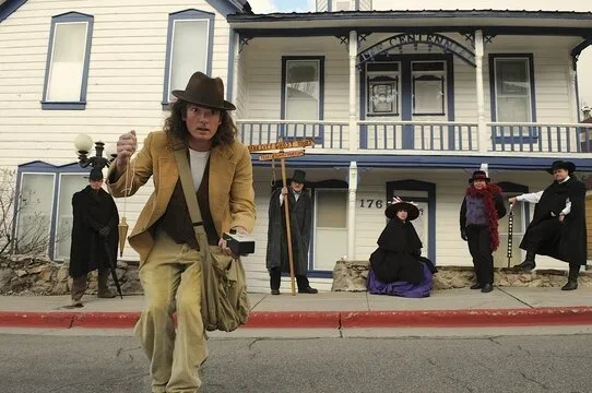 A man in a brown hat and tan jacket walking on the street, with a historic building and four people dressed in old-fashioned costumes in the background.
