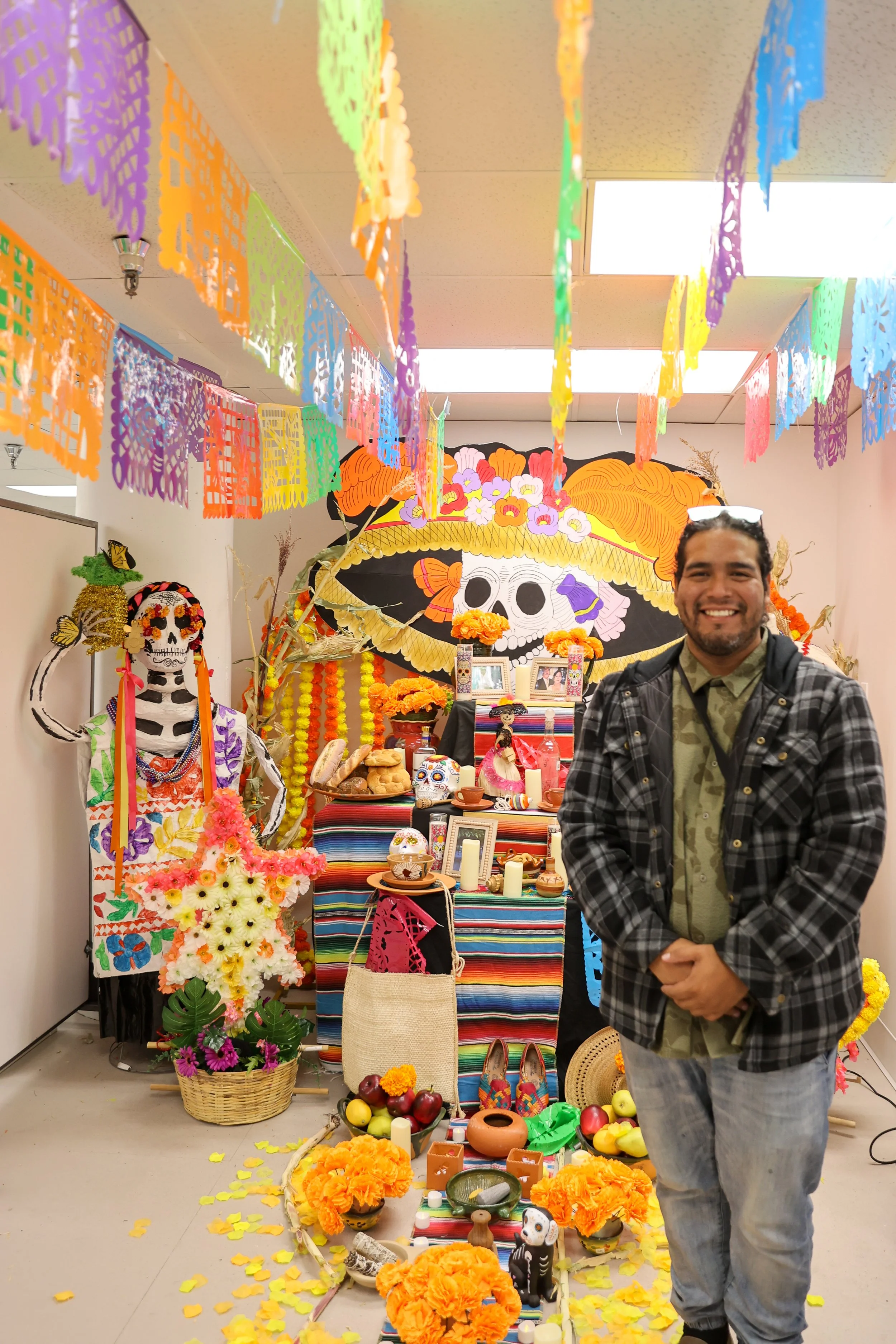 Um homem sorrindo em frente a um altar decorado para o Día de los Muertos, com imagens de caveiras, flores de calêndula, velas, fotos e bandeiras coloridas de papel picado penduradas no teto.