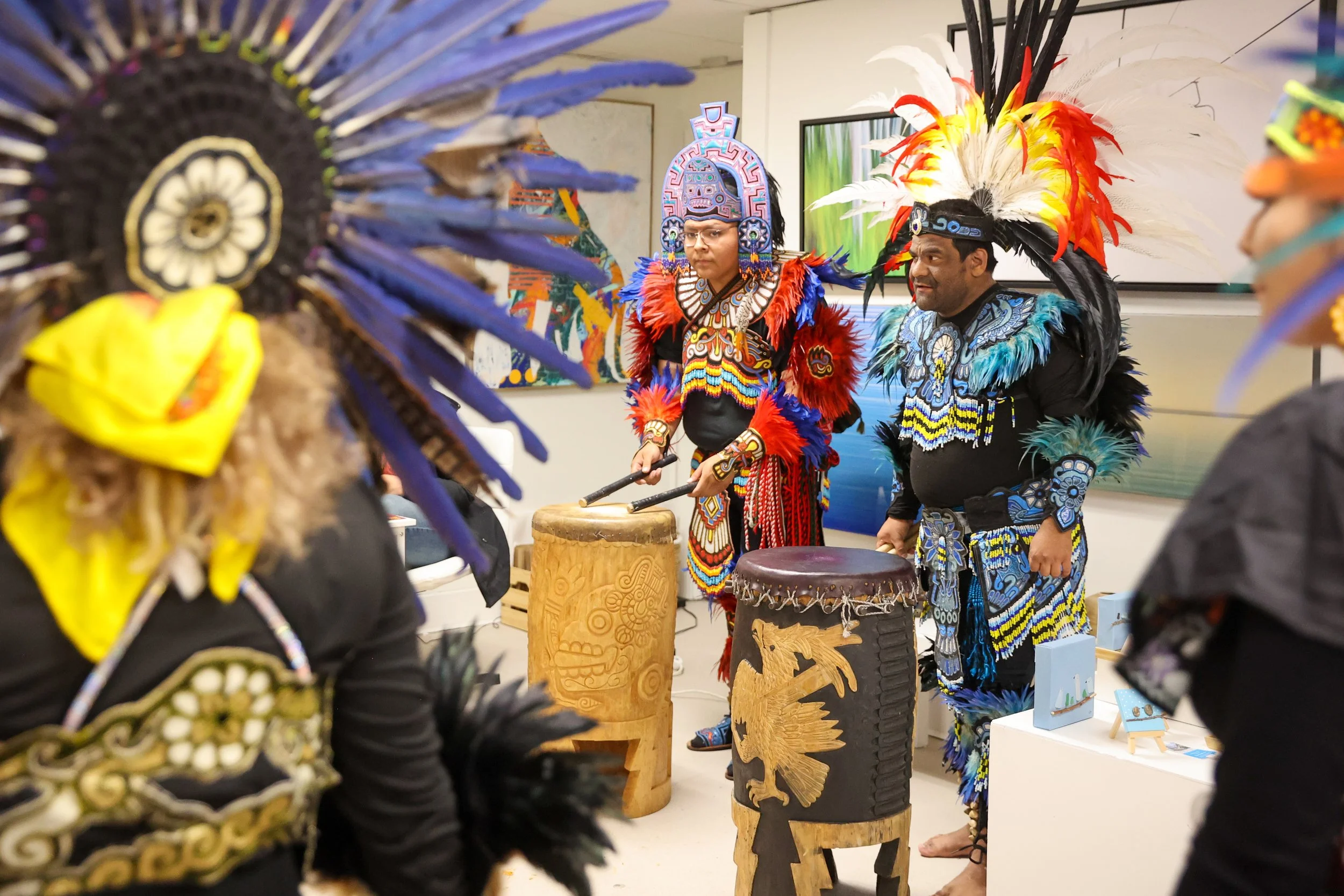 Pessoas vestidas com trajes tradicionais astecas coloridos participando de um evento cultural ou apresentação, com tambores e obras de arte ao fundo.