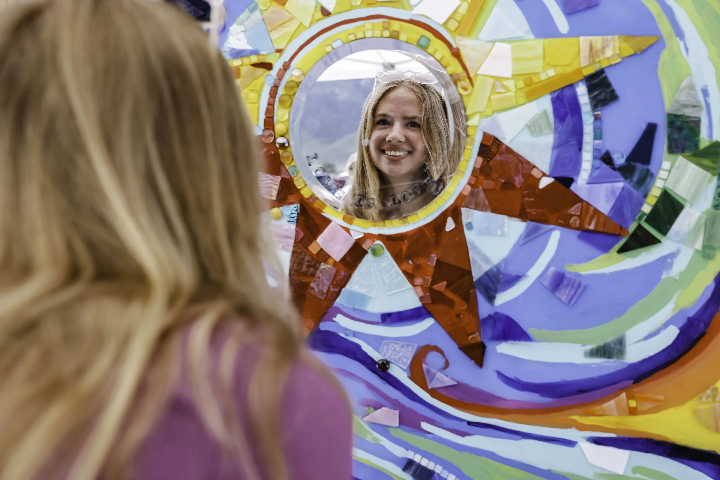A young woman with blonde hair and glasses on her head looking at her reflection in a colorful, mosaic mirror.