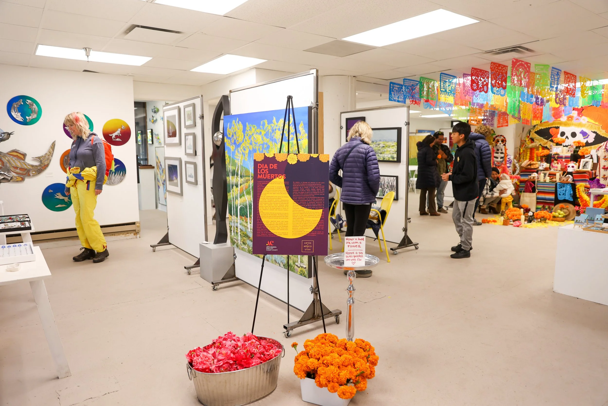 Uma galeria de arte ou mercado interno com decorações coloridas e várias obras de arte. Várias pessoas estão a ver peças de arte, com uma mulher de calças amarelas e um suéter cinza à esquerda, e outras a examinar obras de arte emolduradas nas paredes. Há pa vibrantes