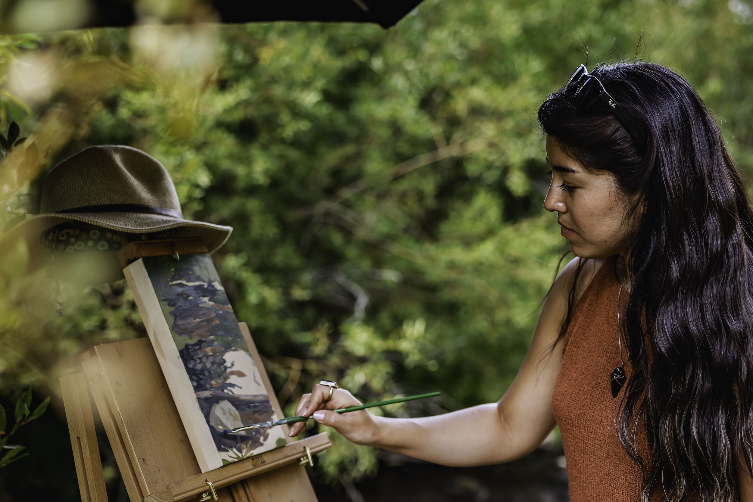 A woman painting outdoors on an easel in a green, wooded area, with a hat resting on the easel.