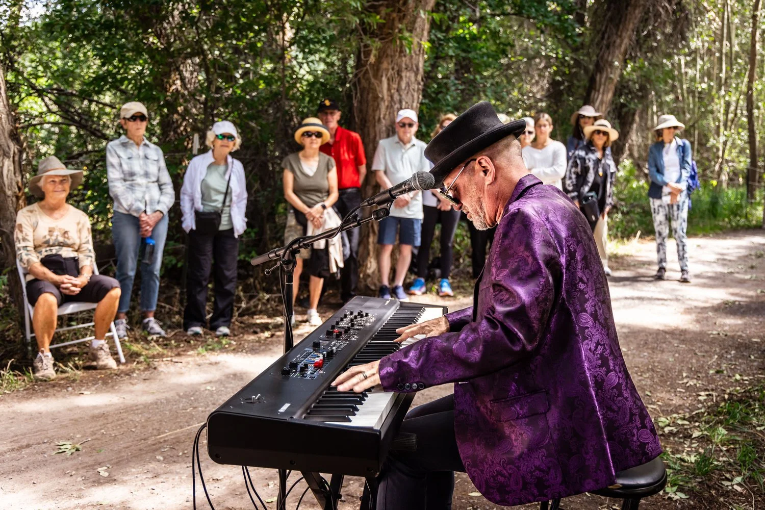 A man wearing a purple blazer and black top hat playing a keyboard outdoors in front of a group of people in a wooded area.