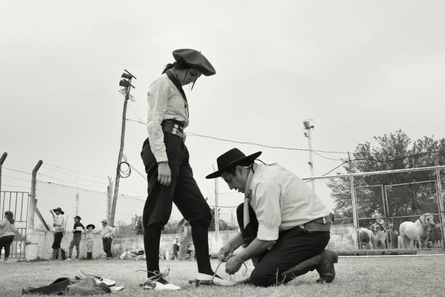 A person dressed as a cowgirl with a hat and Western-style clothing stands in a field while another person kneels tying their shoelaces or adjusting their footwear. Several people and horses are visible behind a fence in the background.