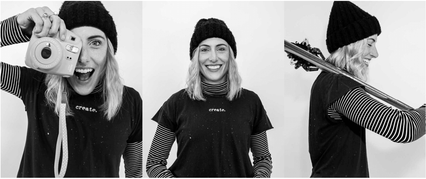 Black and white photo of a woman with blonde hair wearing a black knit beanie, striped long sleeve shirt, and black t-shirt with "create." written on it, posing with a camera and a snowboard.
