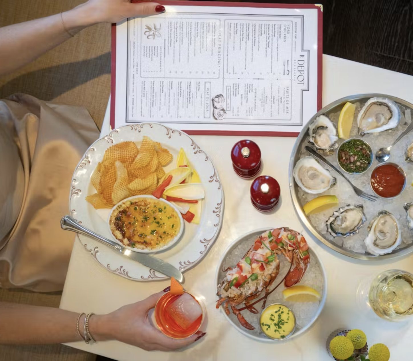 Table with a seafood meal including oysters on a tray with lemon wedges, a bowl of dipping sauce, a plate of scalloped potatoes and salad, and a glass of orange drink. A person is holding the drink.