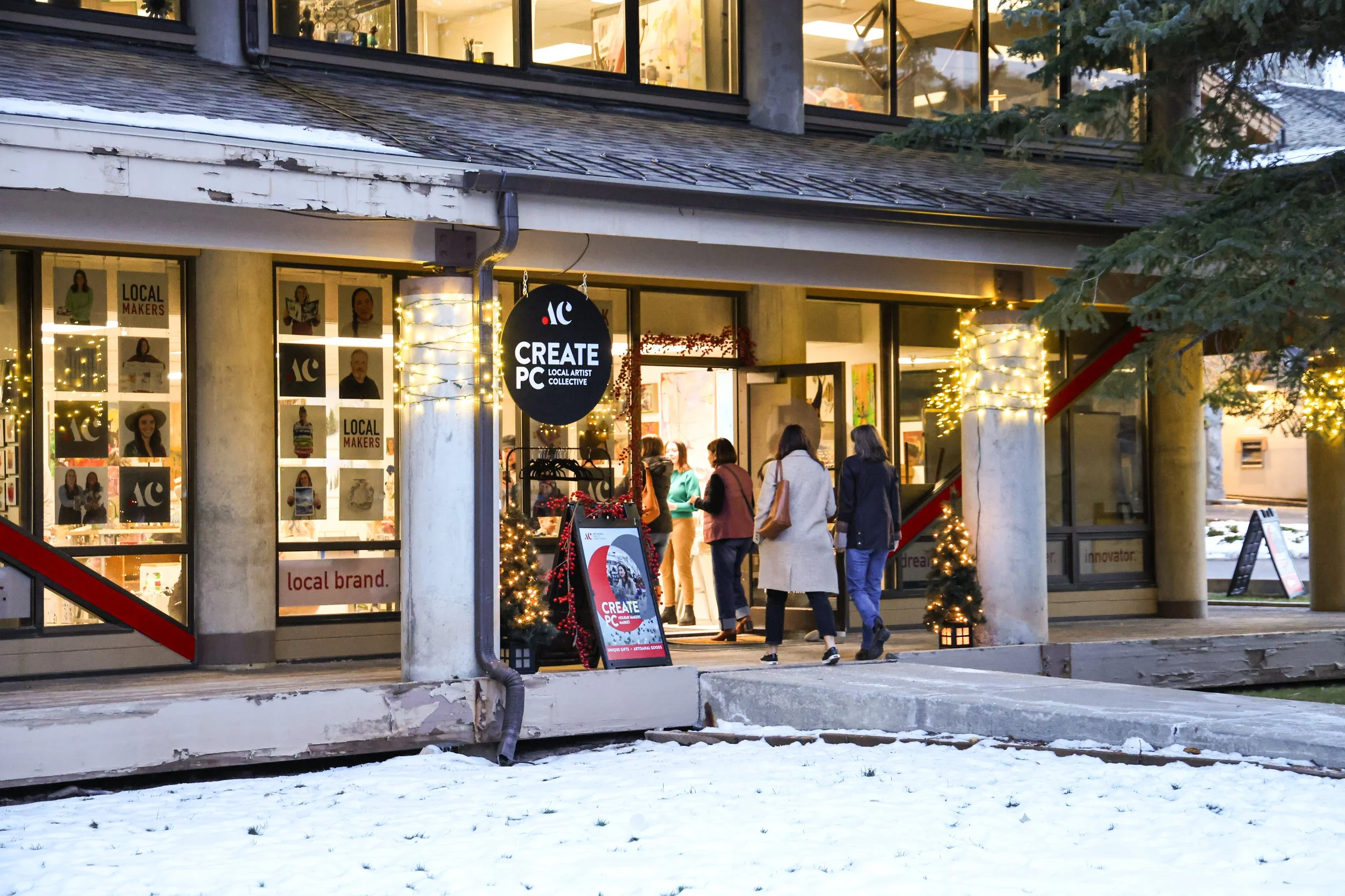 People entering a store decorated with Christmas lights and small Christmas trees in a snowy outdoor setting, with a sign that reads 'Create PC Local Artist Collective'.