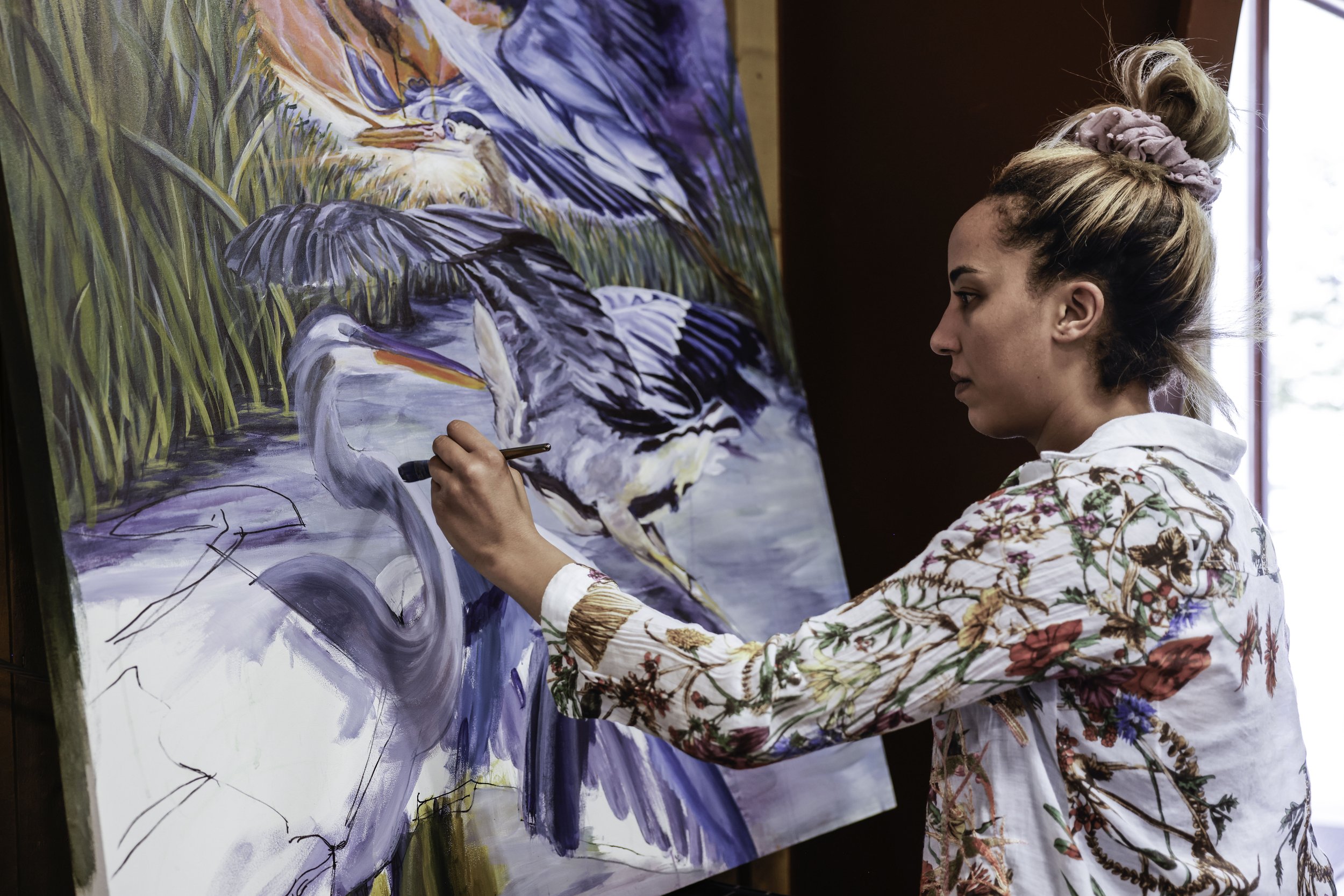 A woman painting a scene with two herons near water and tall grasses on a large canvas in an artist's studio.