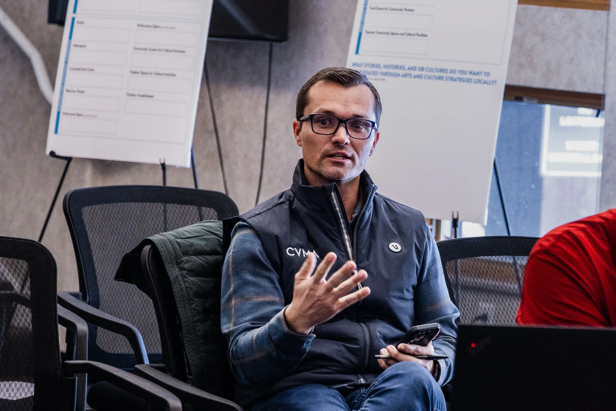 A man with glasses and short dark hair, wearing a black vest and gray jacket, holding a phone, sitting in a chair at a conference or meeting, with whiteboards or presentation boards in the background.