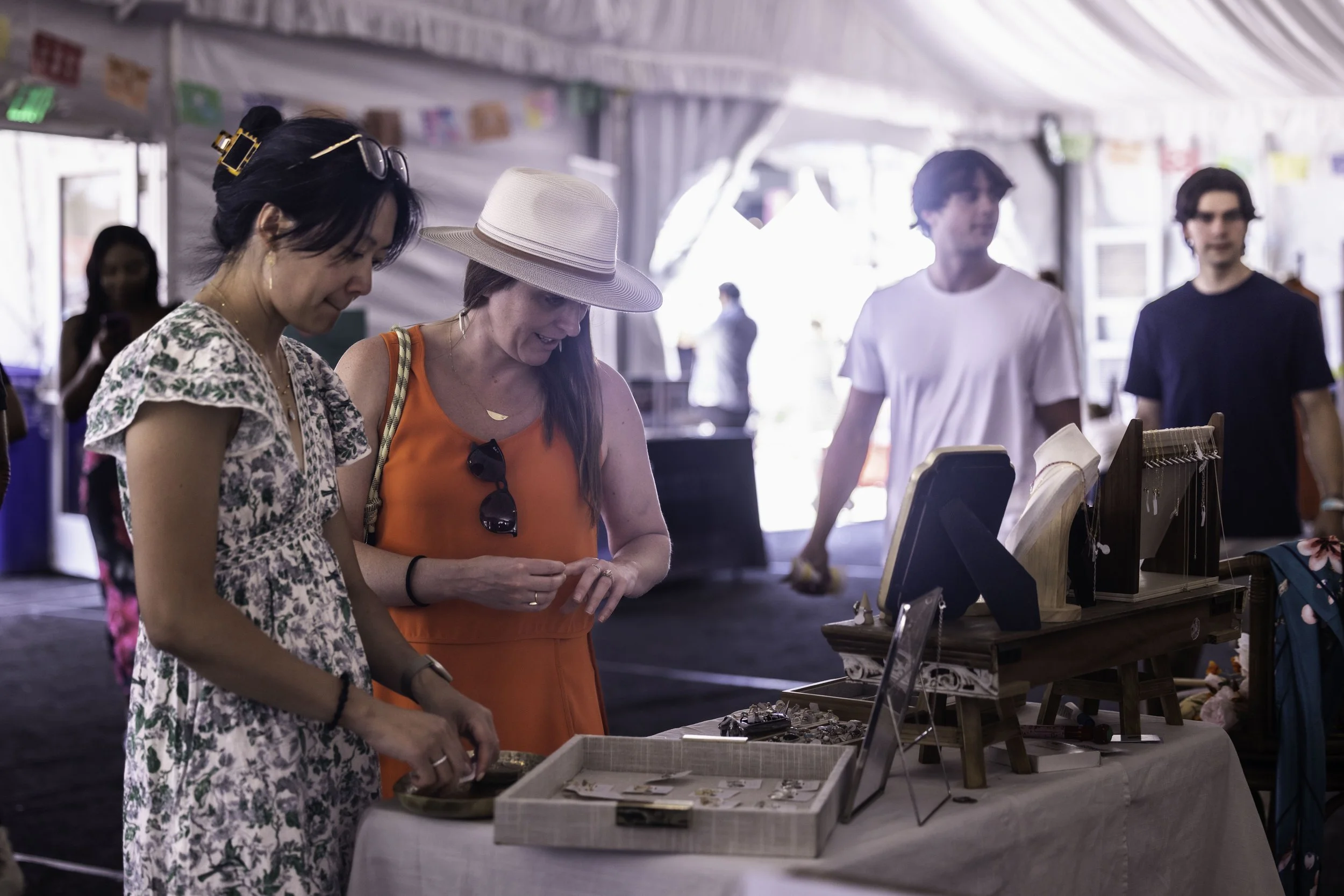Two women browsing jewelry at a craft market stall inside a large tent, with other people and display tables in the background.