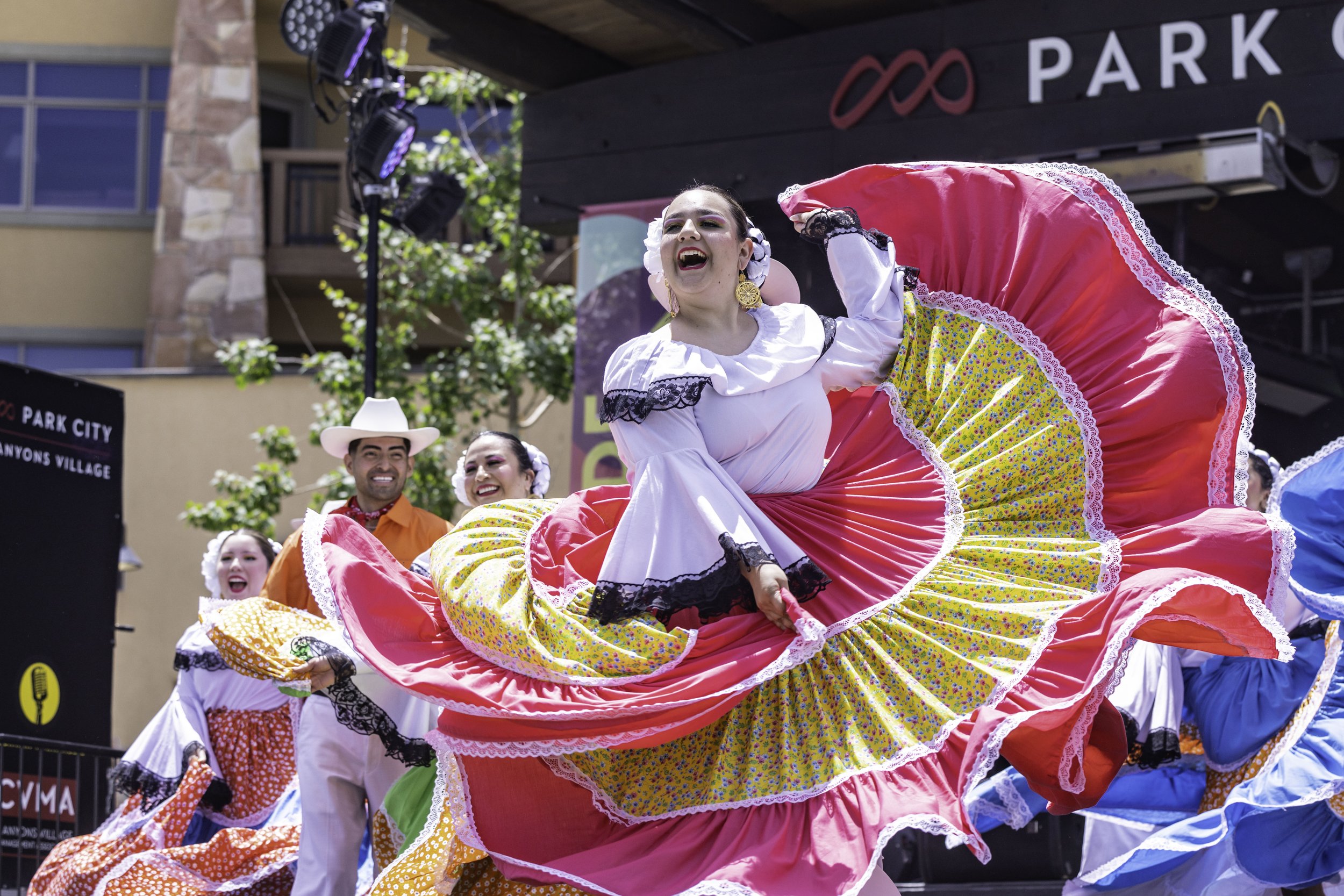 Performers in colorful traditional Mexican dresses dancing outdoors, smiling, during a cultural celebration with a backdrop of buildings and advertising signs.
