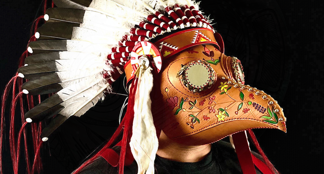 Person wearing a decorative bird mask with floral carvings, feathers, red accents, and a painted faceplate, partially visible against a dark background.