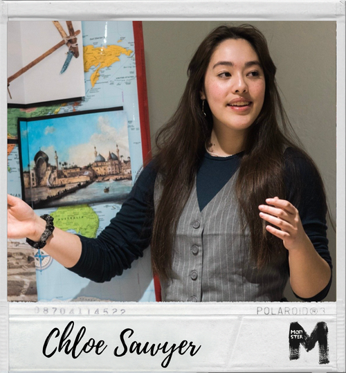 Young woman with long dark hair speaking or explaining in front of a world map and a picture of a castle, wearing a dark sweater and a gray vest.