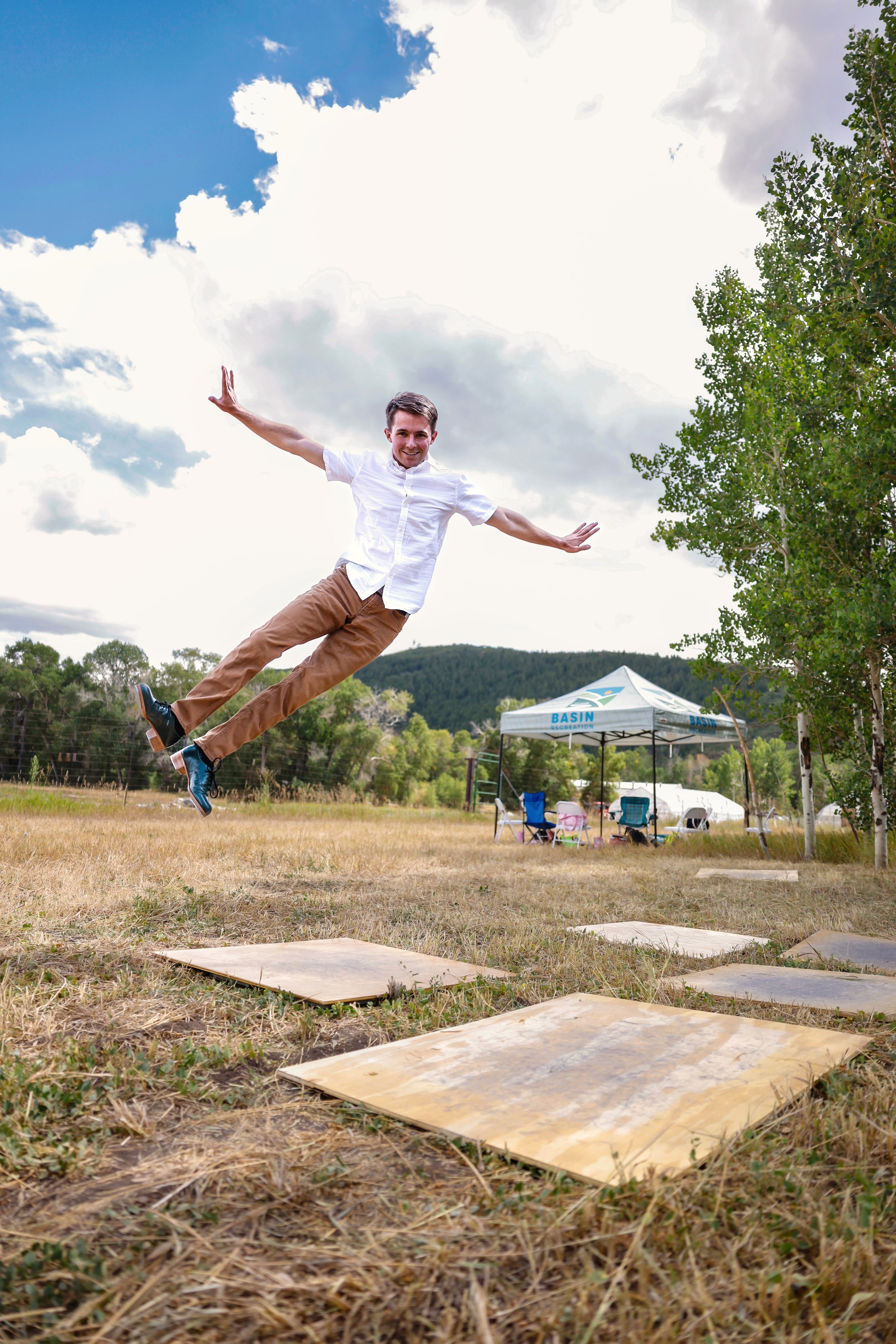 A man in a white shirt and khaki pants jumping on a grassy field with a cloudy sky and trees in the background.