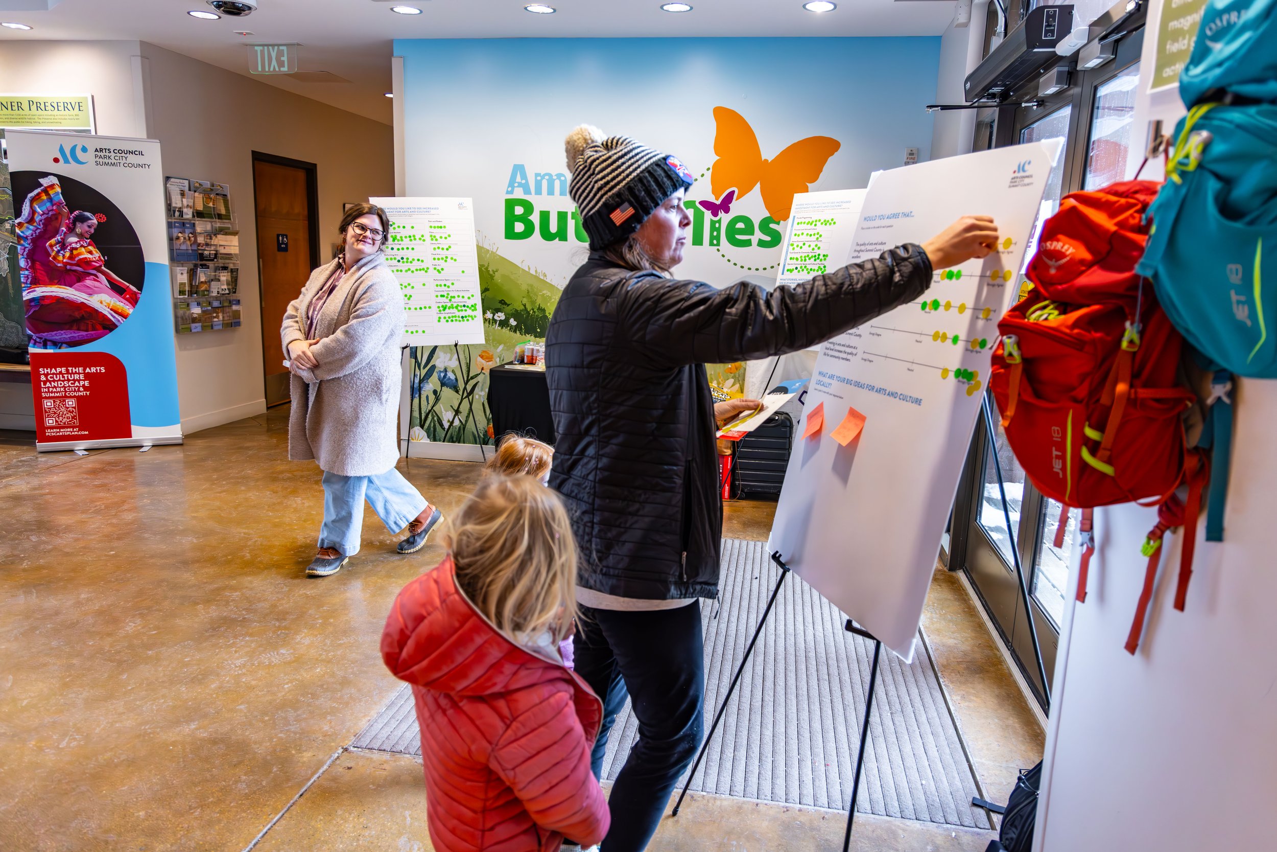 People attending a community event or workshop in a brightly decorated indoor space. Two women, one with a beanie and jacket, are engaging with a large whiteboard, possibly presenting or explaining something.  The background features colorful banners