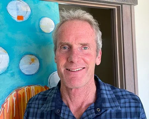 A smiling middle-aged man with gray hair in front of a colorful abstract painting.