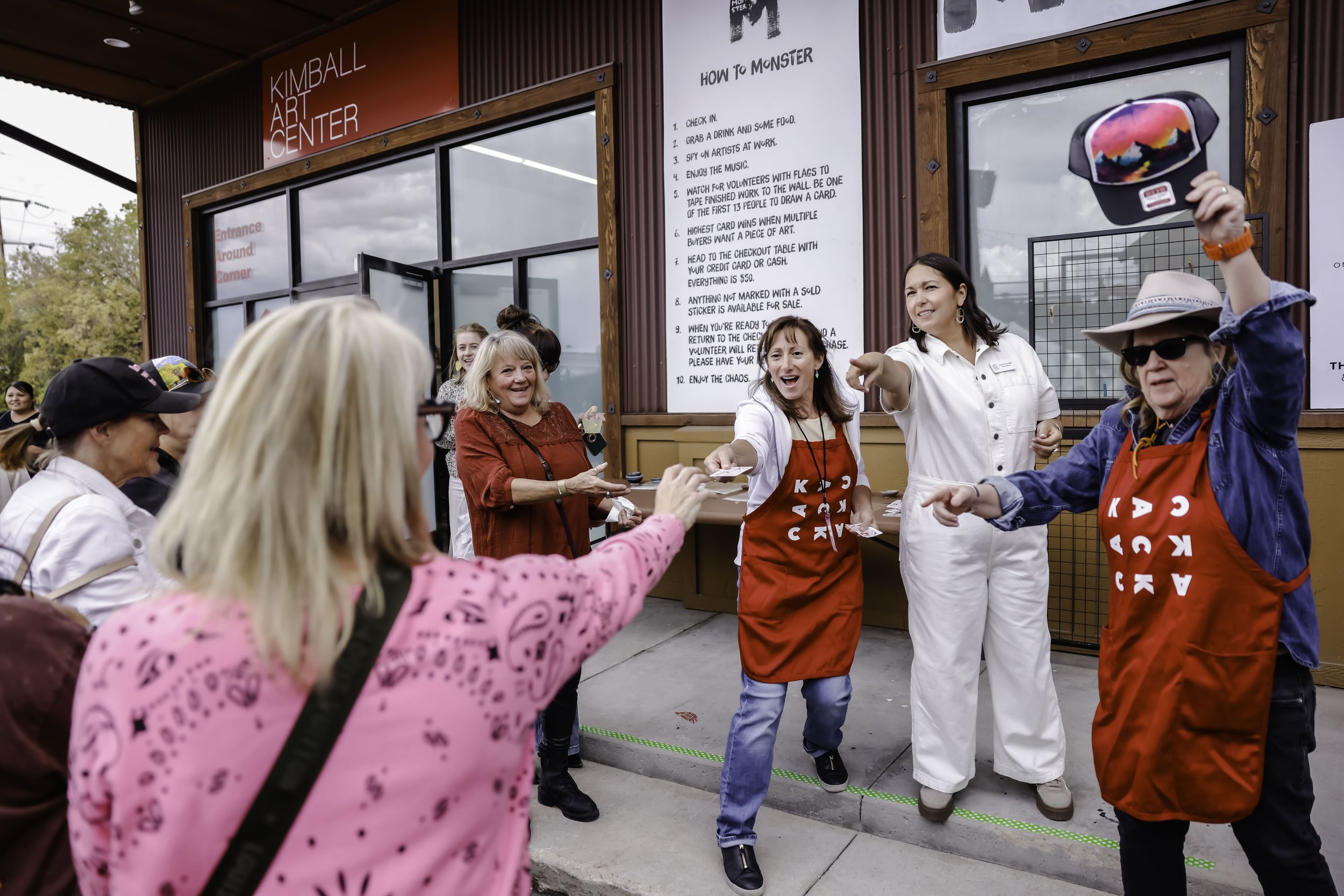 Group of people enjoying a fun activity outside the Kimball Art Center. Two women wearing red aprons are participating in a game while others watch, with one woman pointing and smiling, and a person holding a colorful hat.