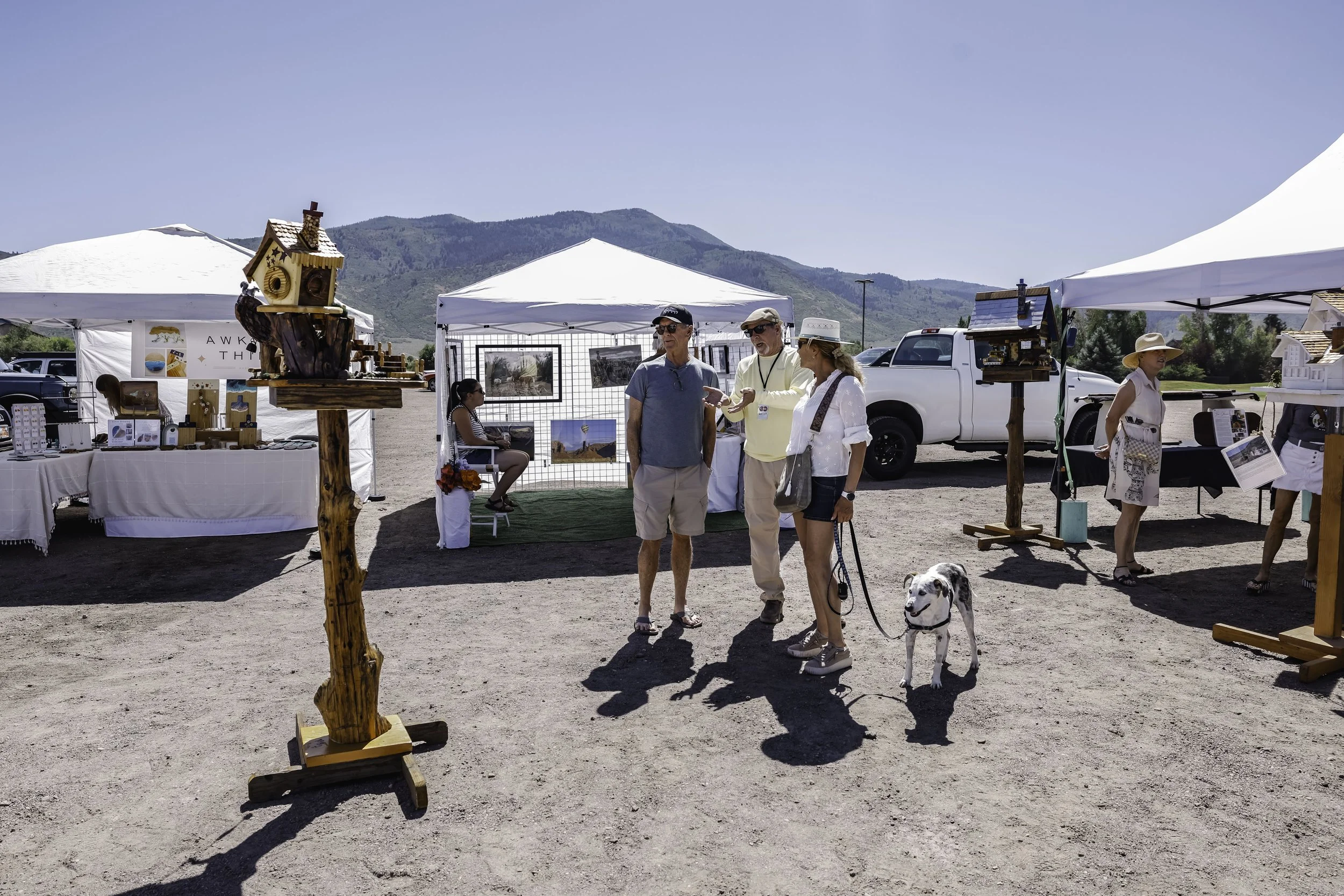 People browsing art and crafts booths at an outdoor market or fair on a sunny day, with mountains in the background.