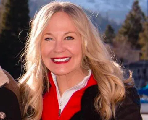 Smiling blonde woman outdoors with trees and mountains in the background.