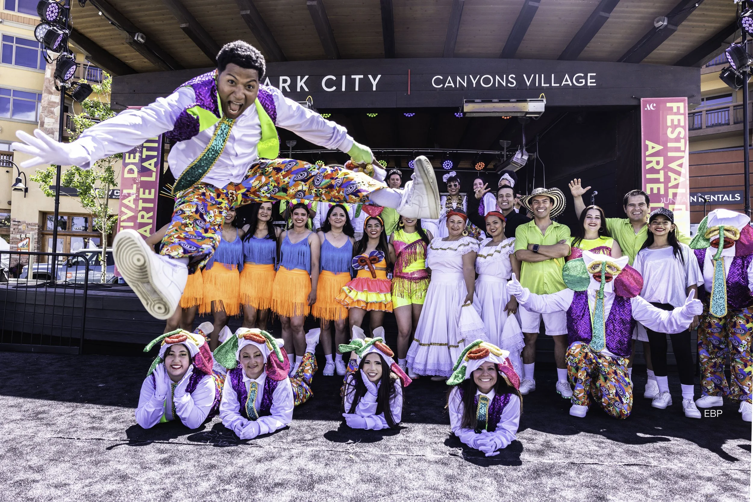 Group of performers dressed in colorful costumes on an outdoor stage, with a man jumping in the foreground. The stage background has signs reading 'Park City' and 'Canyons Village'.
