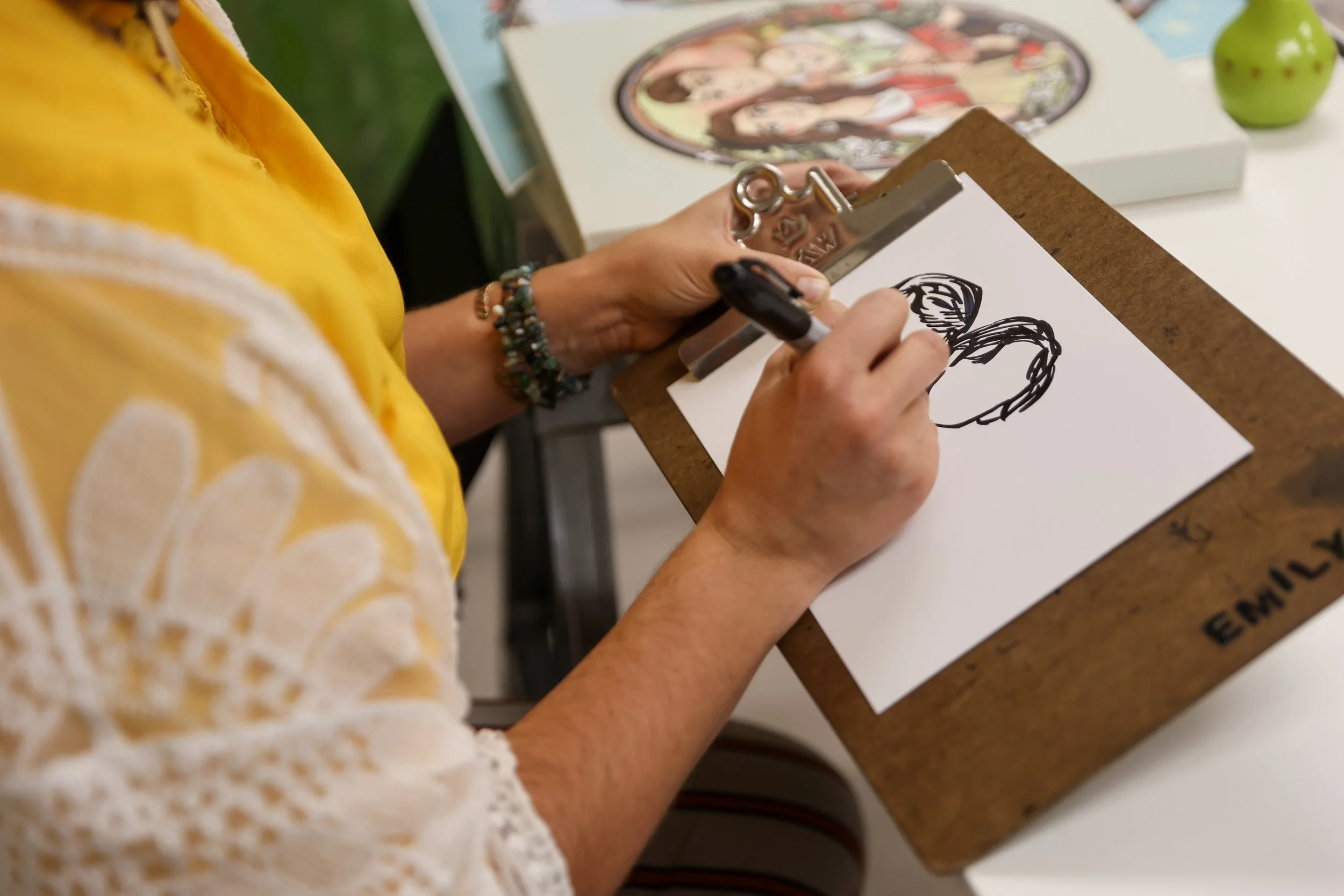 Uma pessoa vestindo uma blusa amarela e branca com ombros à mostra está a desenhar um coração com um marcador preto num pedaço de papel branco preso a uma prancheta marrom, sobre uma mesa. Ao fundo, há livros e uma maçã verde.