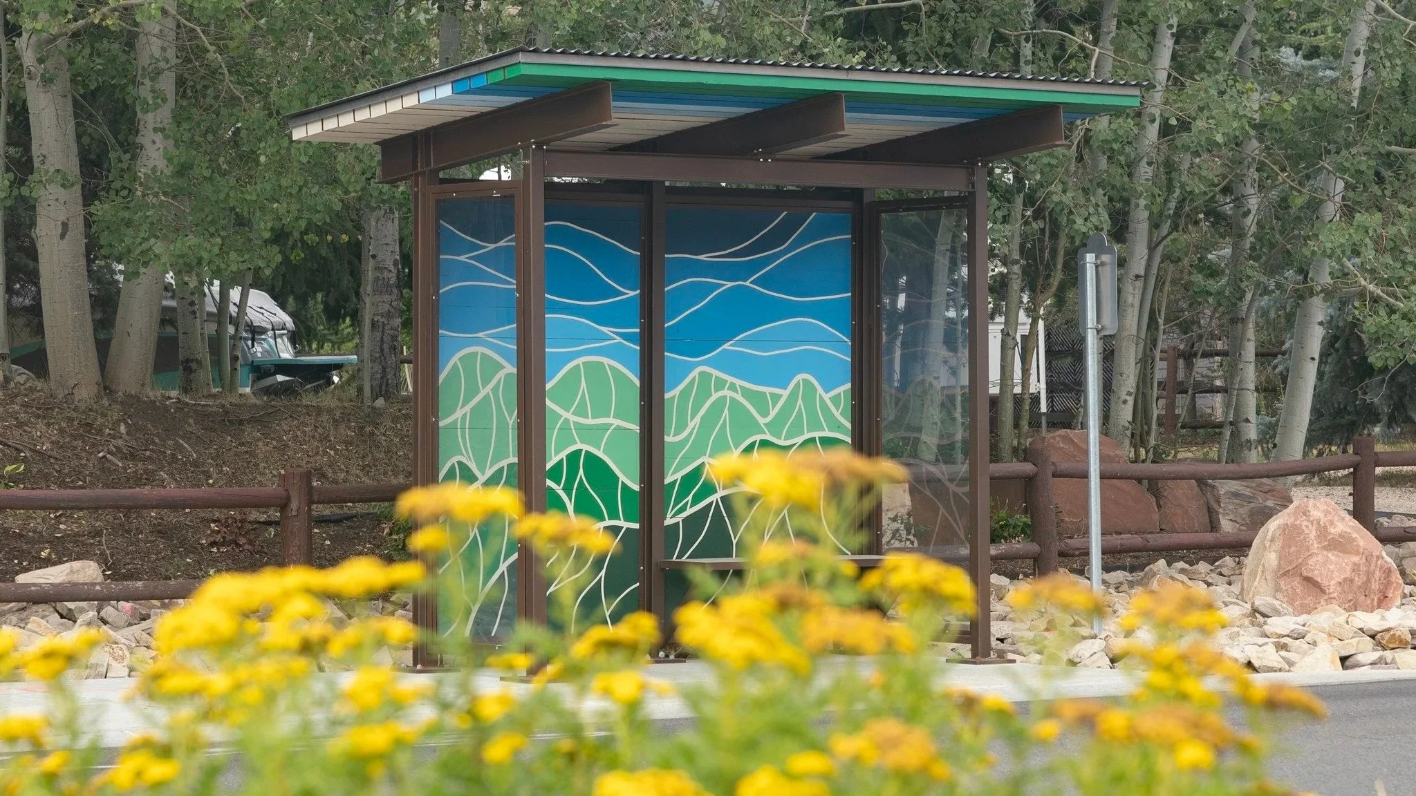A bus stop shelter with colorful mural art featuring rolling green hills and blue sky, located along a roadside with yellow flowers in the foreground.