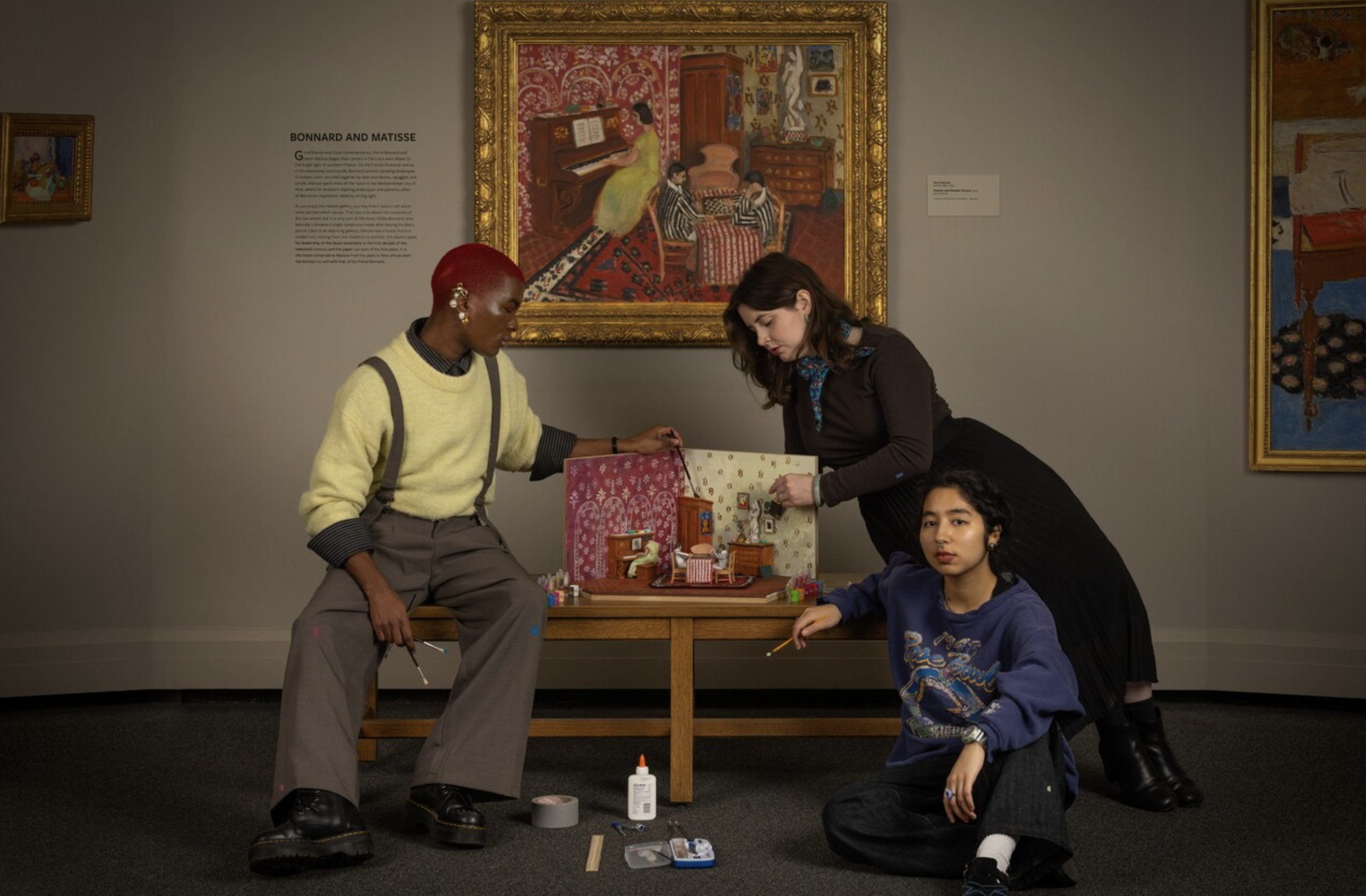 Three women in an art gallery working on a dollhouse or miniature room display. One woman with red hair and yellow sweater is on the left, another with dark hair and dark clothing leans over the display in the center, and a woman with curly hair in a blue sweatshirt sits on the floor on the right. The gallery walls have framed paintings.