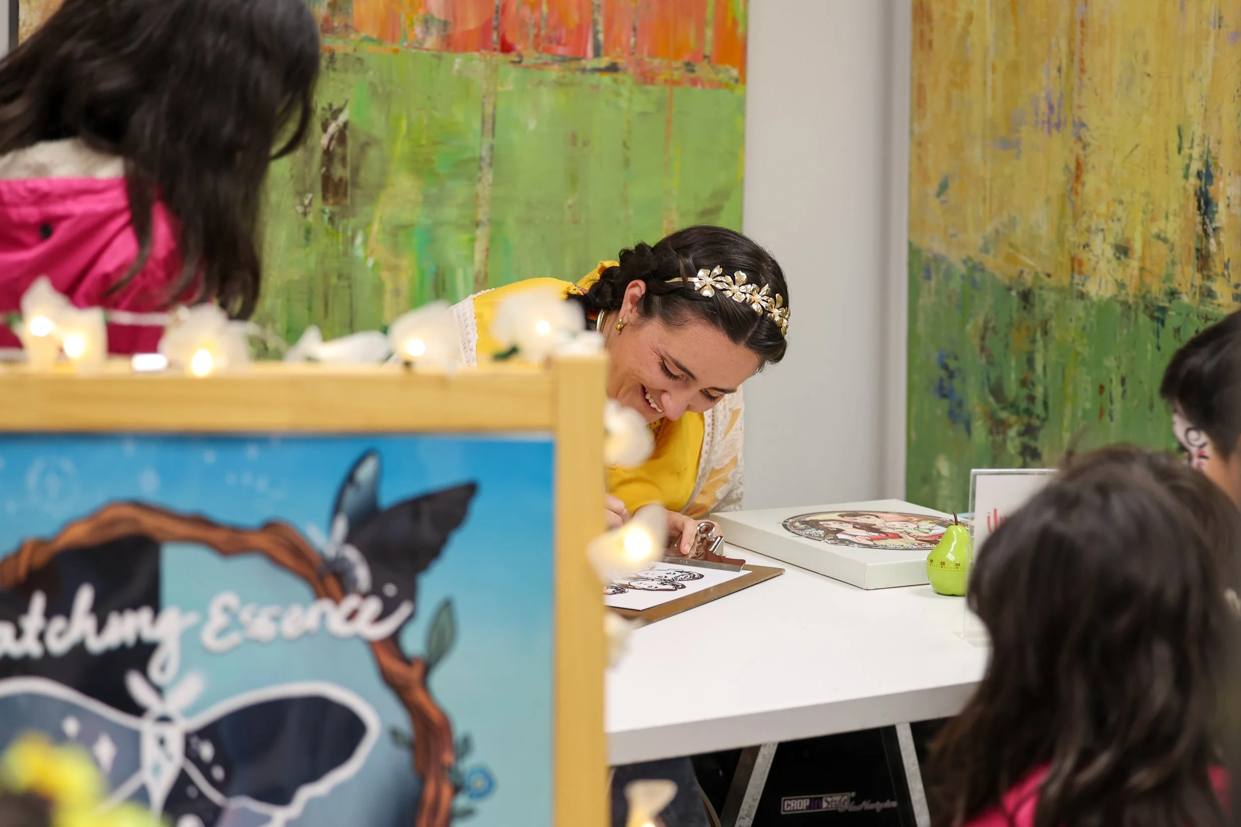 Uma mulher com uma fita floral na cabeça e uma camisa amarela sorrindo e a dar autógrafos num evento de lançamento de livro, rodeada por crianças e obras de arte coloridas ao fundo.