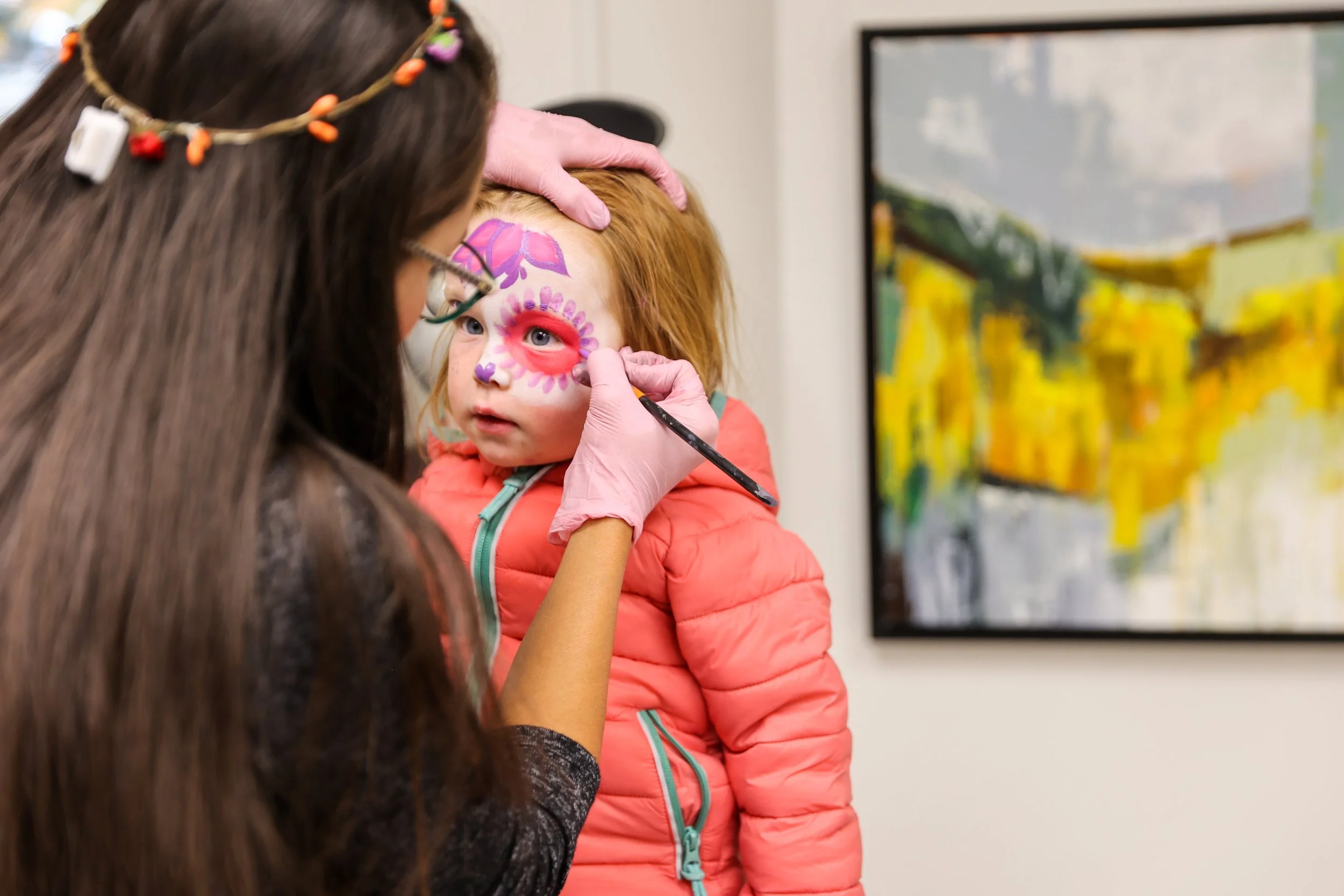 Uma jovem com pintura facial sendo pintada por um artista, com uma pintura colorida ao fundo.