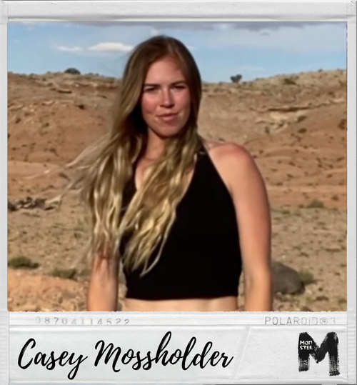A young woman with long, wavy blonde hair wearing a black sleeveless top outdoors in a desert landscape with rocks and sparse vegetation, under a partly cloudy sky.