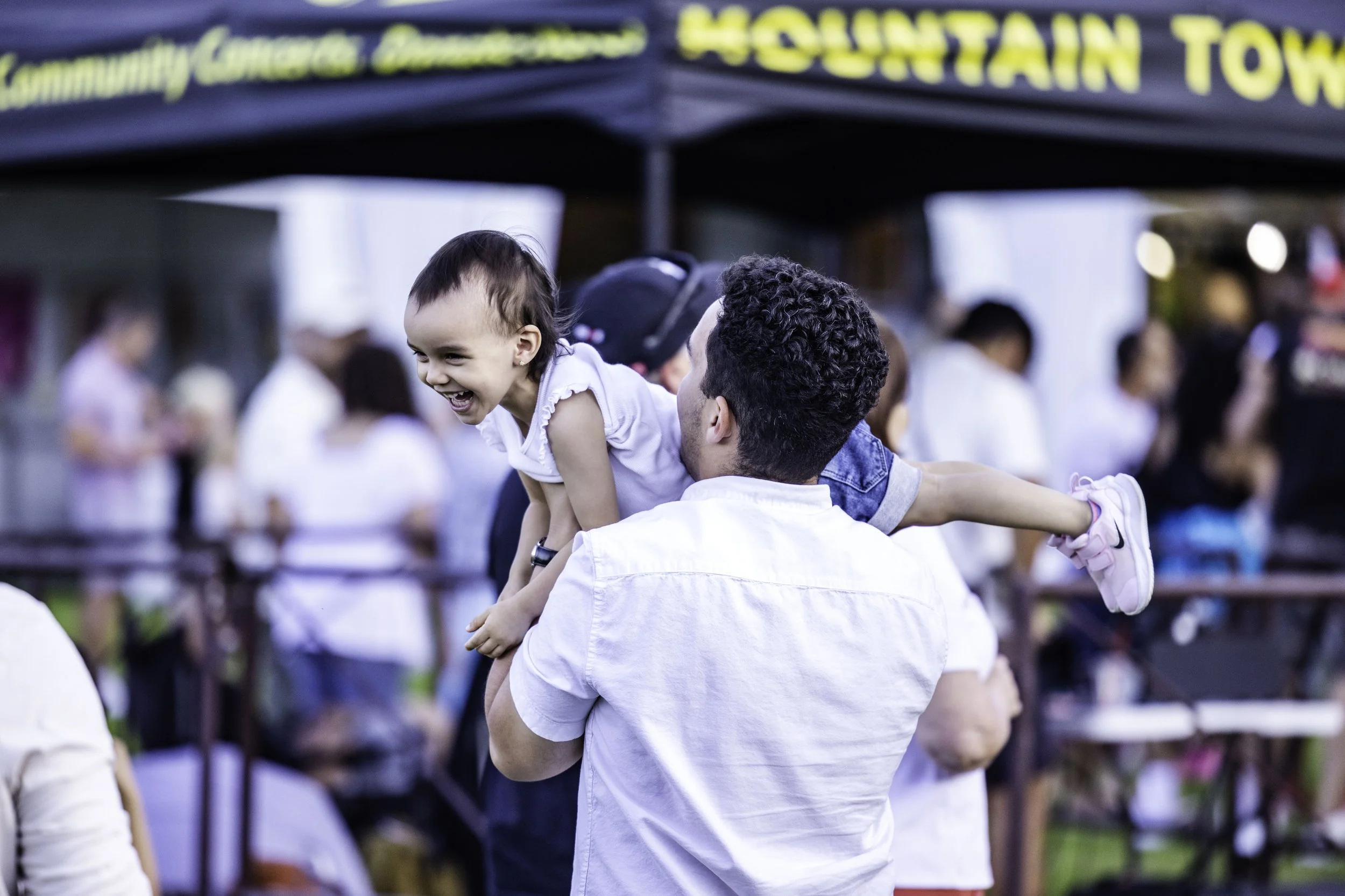 A man is holding a smiling young girl on his shoulders at an outdoor event. The girl is laughing and wearing a white shirt and pink sneakers. There are people in the background near a booth with a sign that reads 'Mountain Town'.