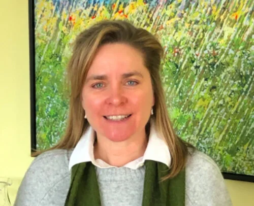 A woman with light brown hair smiling in front of a colorful abstract painting.