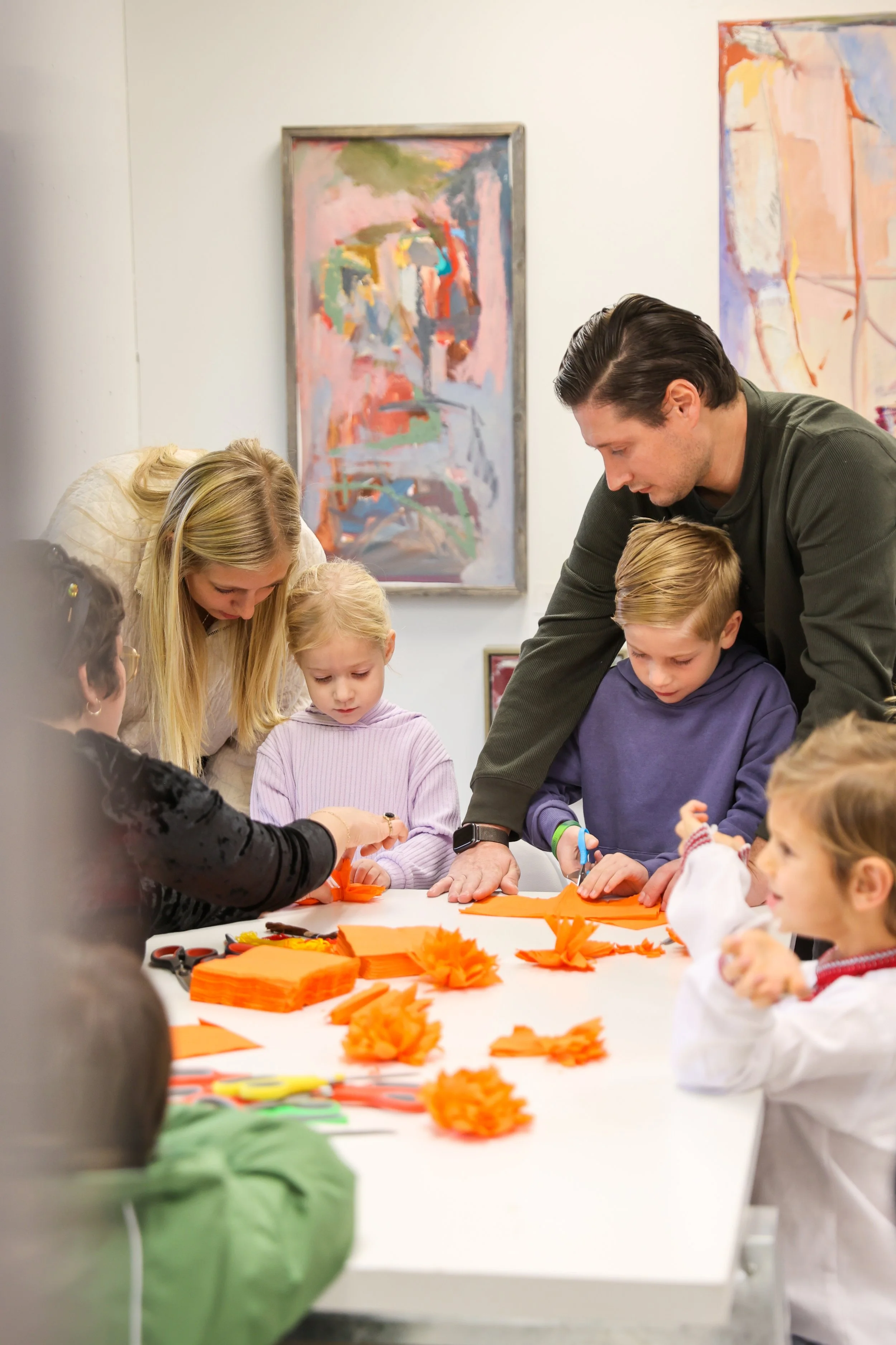 Crianças e adultos reuniram-se em torno de uma mesa para criar flores de origami com papel laranja numa galeria de arte.