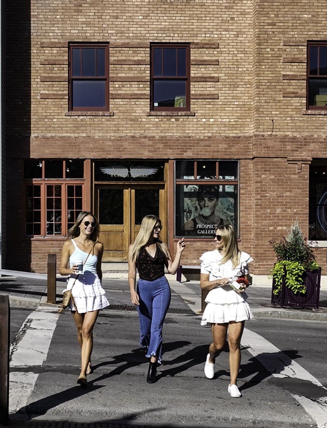 Three young women walking across a crosswalk in front of a red brick building with large windows, with one woman holding a drink, another holding a phone, and the third carrying a small object.