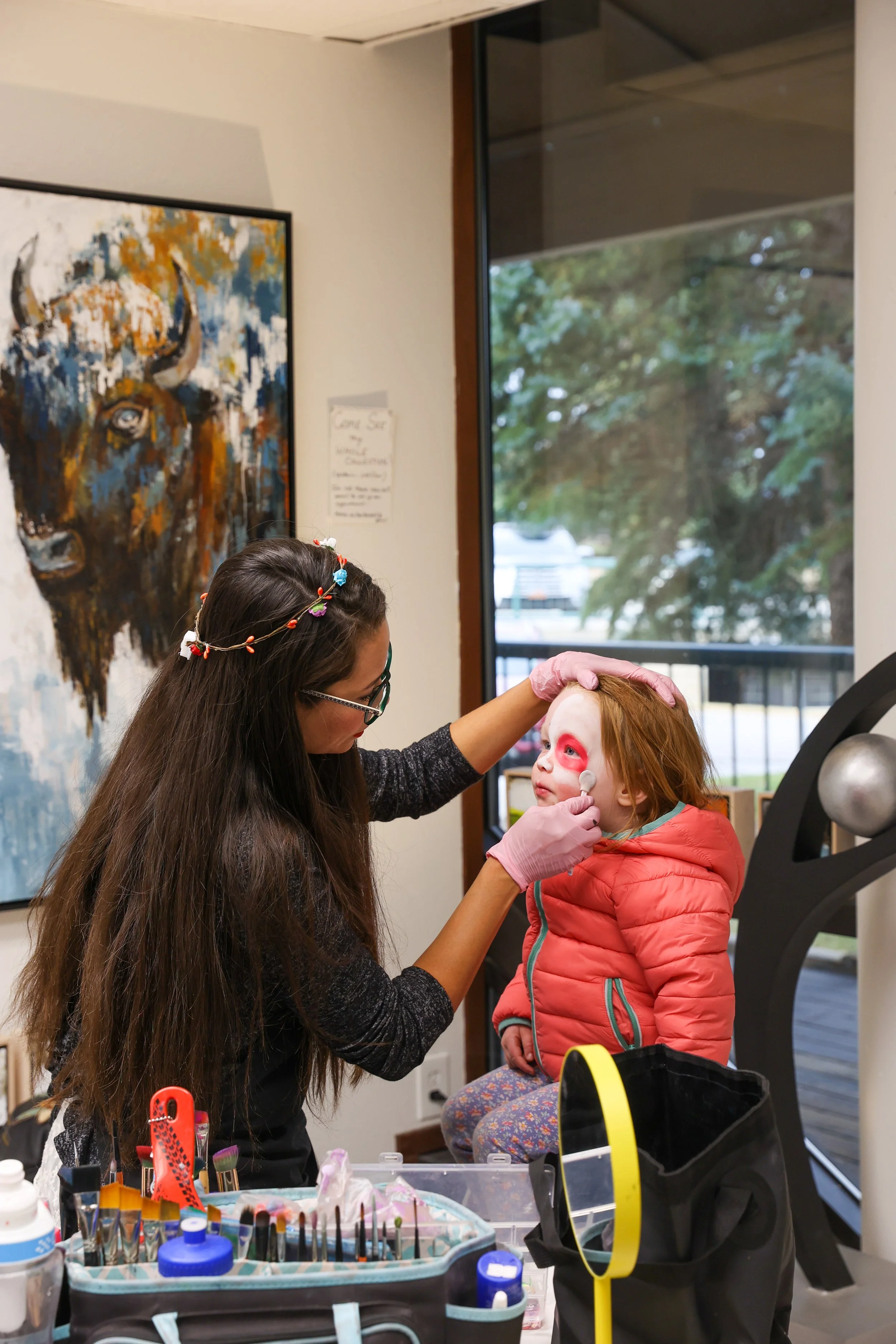 Uma mulher a pintar o rosto de uma menina sentada numa cadeira, com materiais para pintura facial numa mesa próxima, dentro de uma sala com uma grande janela e uma pintura de um búfalo na parede.