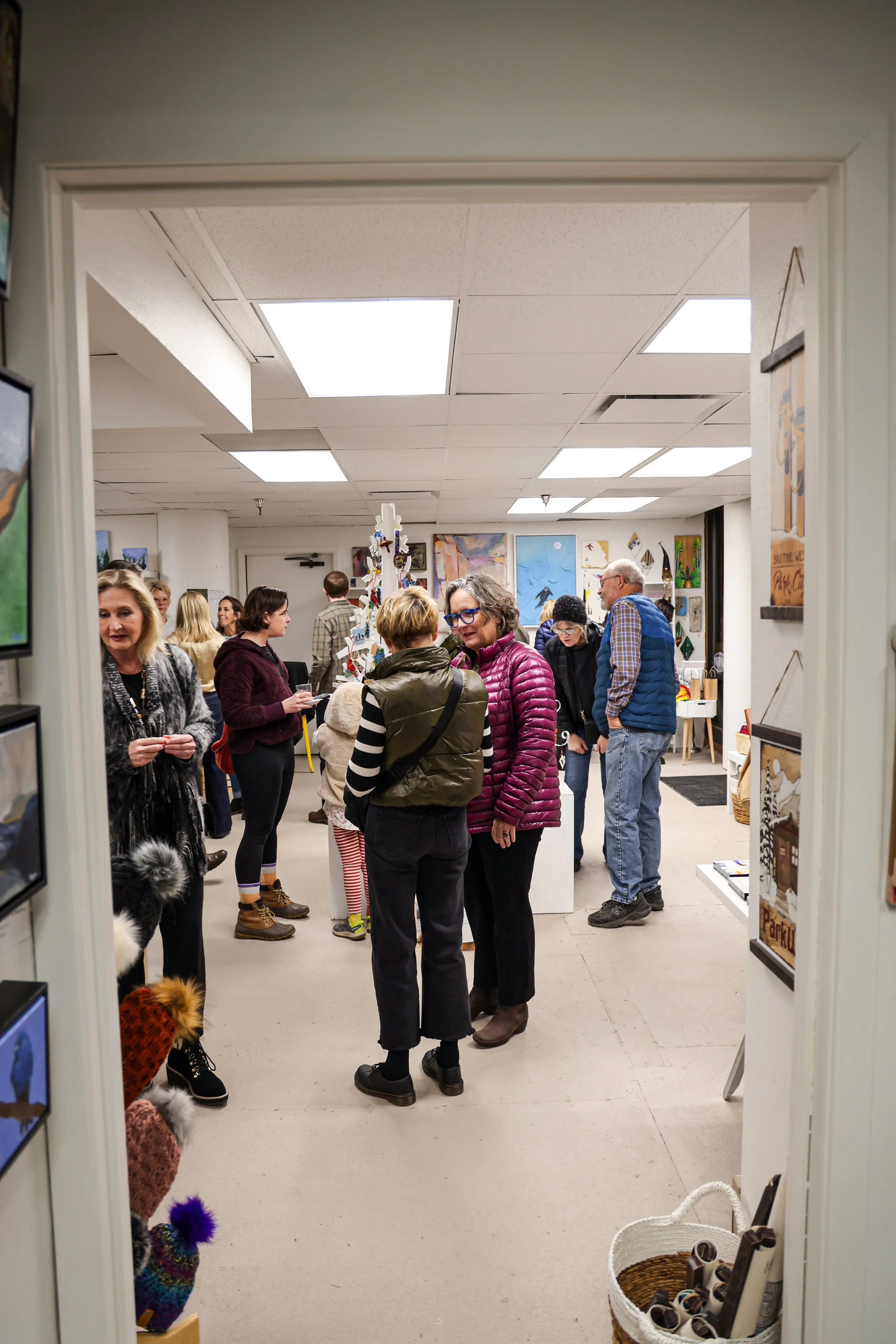 People socializing at an art gallery, with artwork and paintings displayed on walls, and shelves with art supplies nearby.