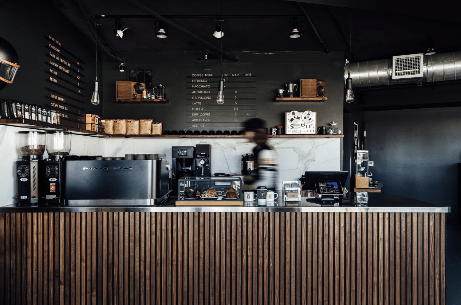 Counter and back wall of a modern coffee shop with coffee menu, espresso machines, baked goods, and a barista in motion.