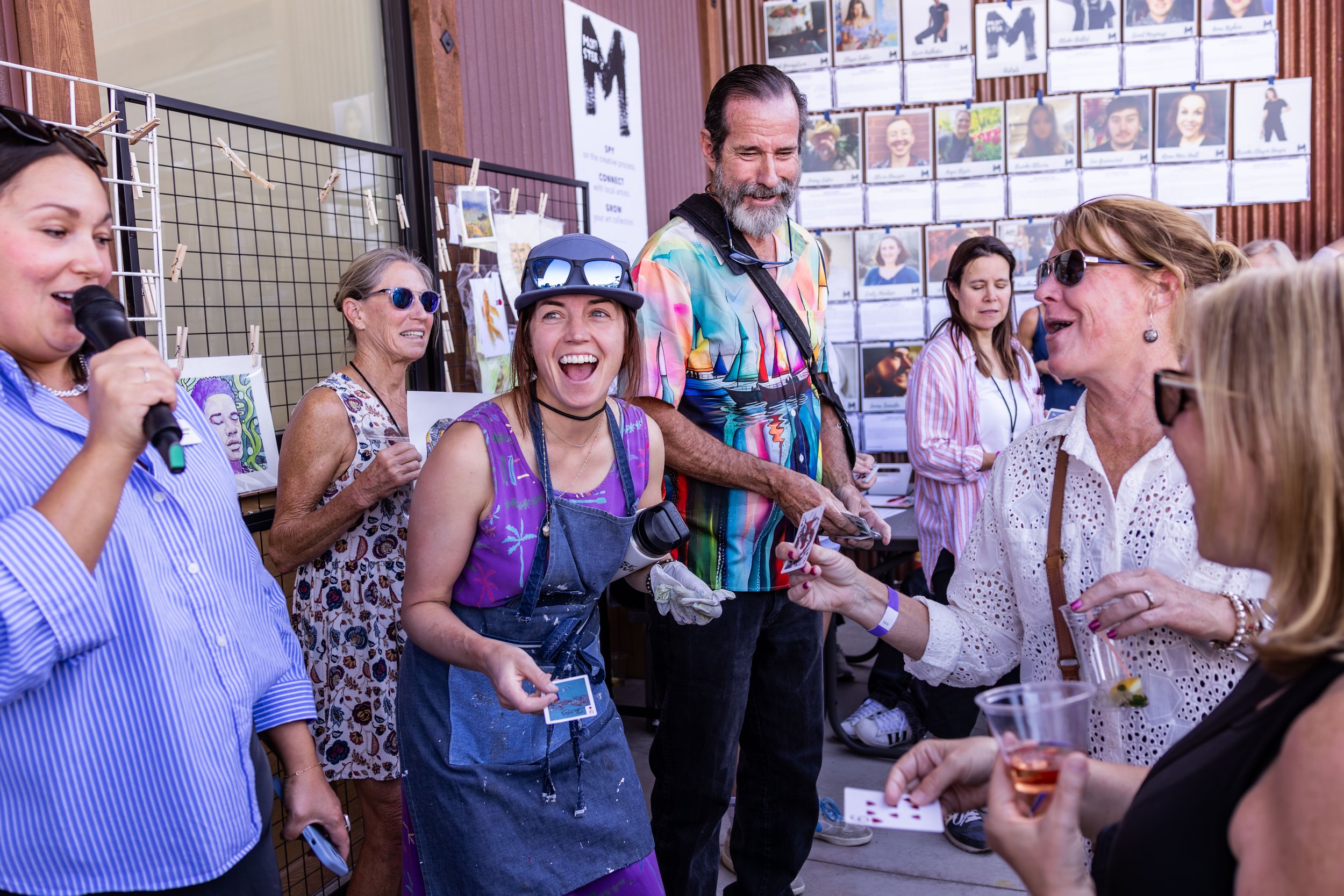 Grupo de pessoas num mercado ou feira de arte ao ar livre, conversando e rindo, com obras de arte expostas nas paredes e mesas atrás delas.