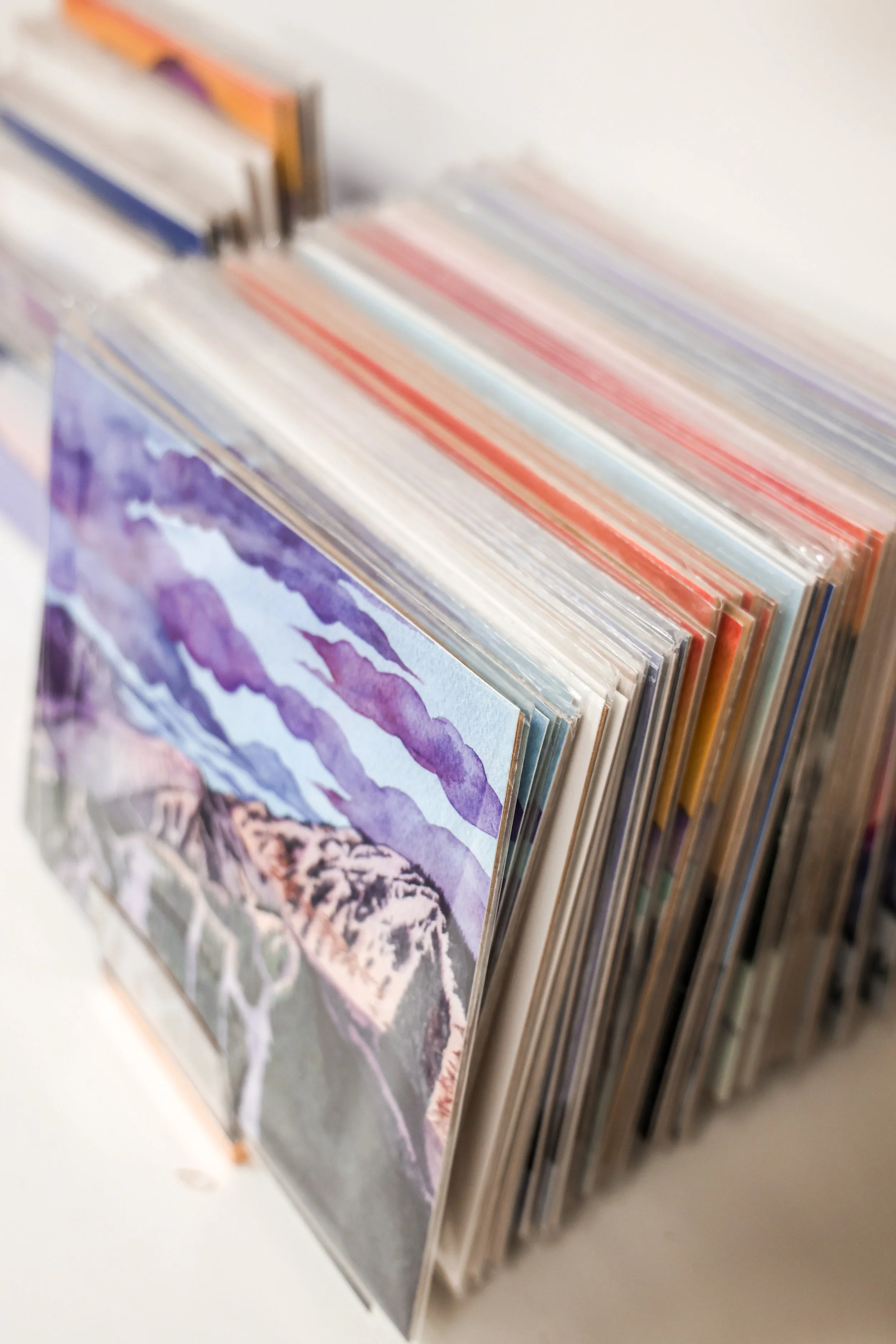 Close-up of a collection of postcards or prints, with one featuring a watercolor painting of mountains, purple sky, and hiking trail in the foreground.