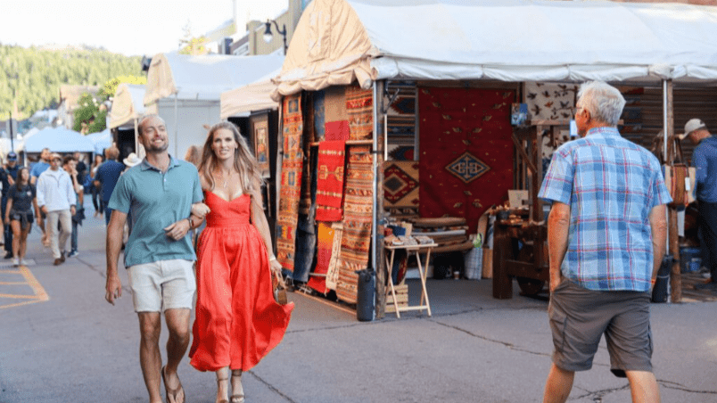 People shopping and walking at an outdoor market with colorful rugs and textiles displayed at vendor stalls.