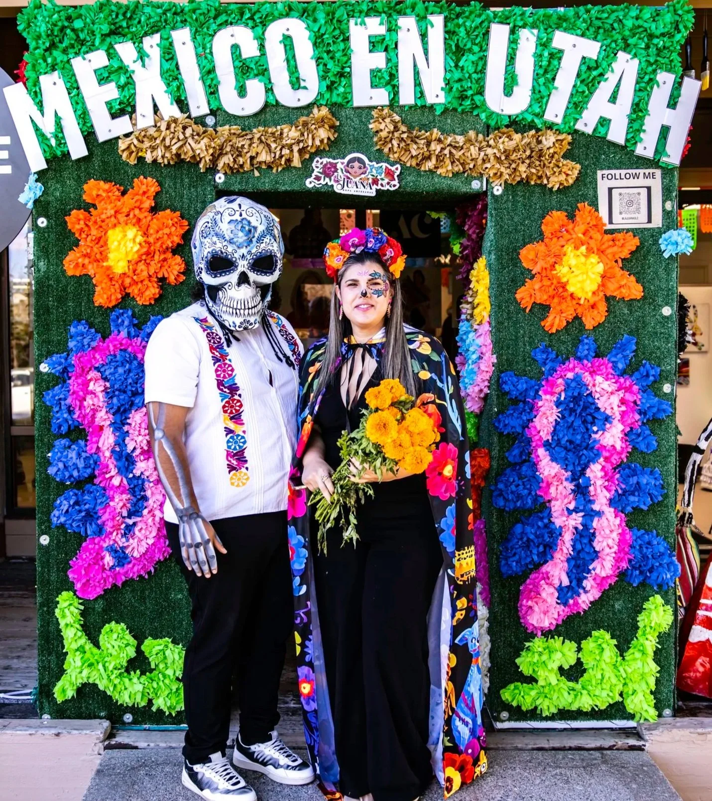 Two people dressed in Día de los Muertos costumes standing in front of a colorful flower and papel flower backdrop with a sign that reads 'MEXICO EN UTAH!' and a QR code.