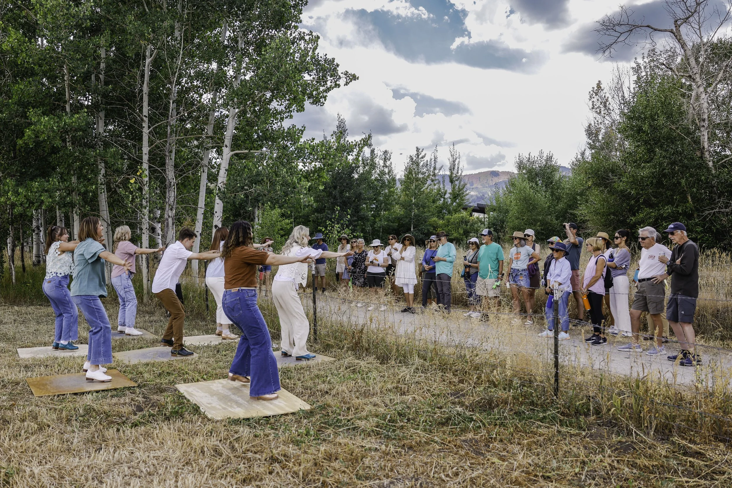 Um grupo de pessoas ao ar livre numa zona rural, a assistir a uma fila de mulheres em plataformas de madeira a dançar descalças, rodeadas por árvores e relva, com montanhas ao fundo.