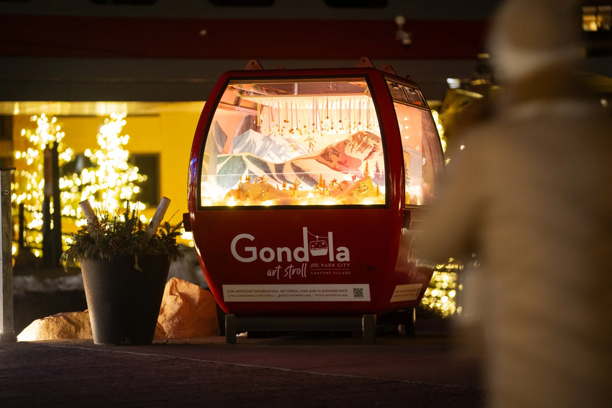 A red gondola-shaped art installation with a miniature mountain scene and festive lights, located outdoors at night. There is a potted plant with pine branches and ornaments nearby, and blurred yellow-lit buildings and trees in the background.