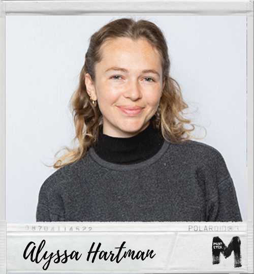 Young woman with curly blonde hair and a black turtleneck under a dark gray sweater, smiling at the camera, against a plain light background.
