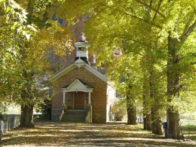 A small brick church with a steeple, surrounded by large trees with green and yellow leaves, and a dirt path leading to its front steps.