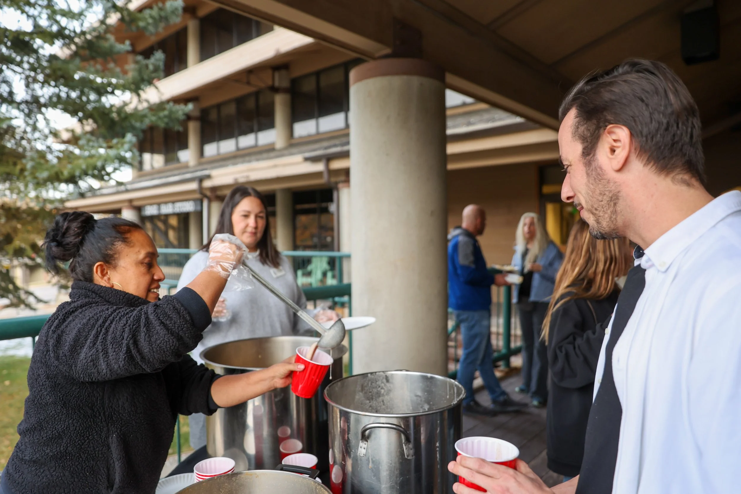 Pessoas servindo e recebendo bebidas quentes em um evento ao ar livre, com uma mulher entregando um copo a um homem e mais pessoas ao fundo.