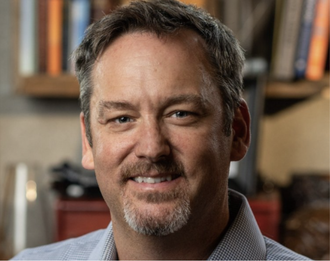 A man with short brown hair and a goatee, smiling at the camera, in front of a blurred background of bookshelves.
