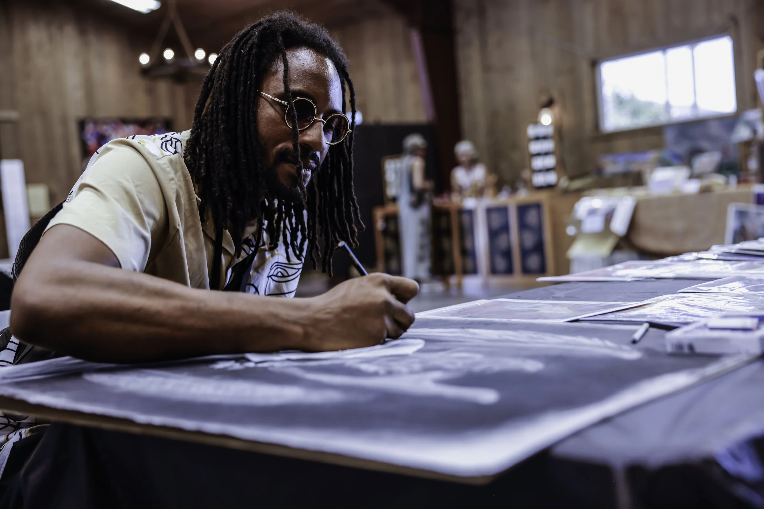 A man with dreadlocks and glasses sitting at a table, drawing on large sheets of paper, in a wooden interior space. In the background, there are people at a counter and a window.