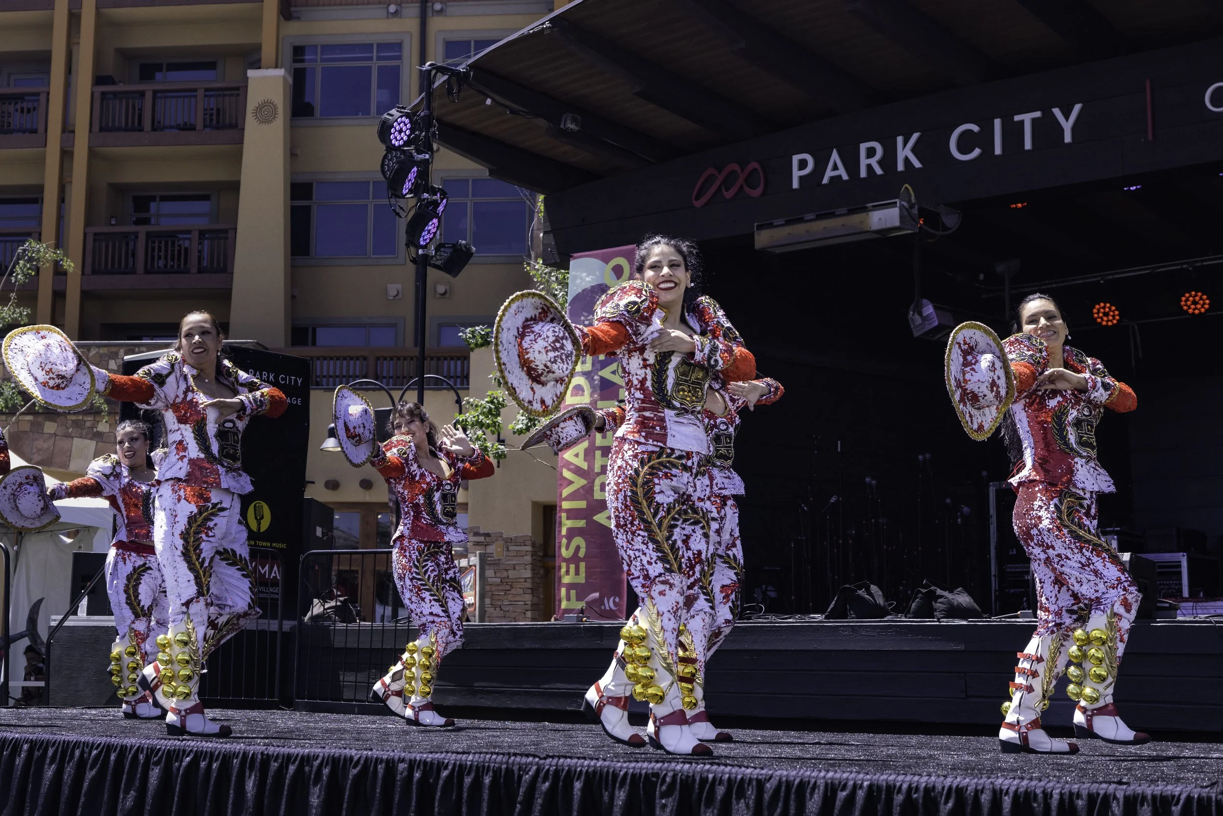 Five women in colorful costumes performing a dance on an outdoor stage at a festival, with buildings in the background.